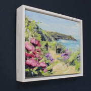 a framed Jill Hudson painting of Rame Head in Cornwall with pink flowers in foreground it is on a dark blue wall