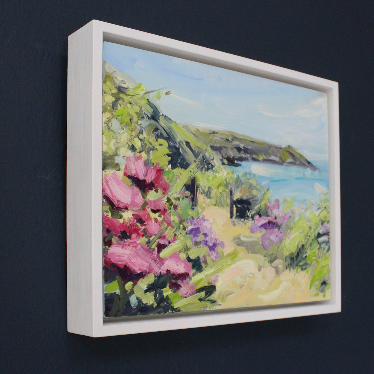 a framed Jill Hudson painting of Rame Head in Cornwall with pink flowers in foreground it is on a dark blue wall