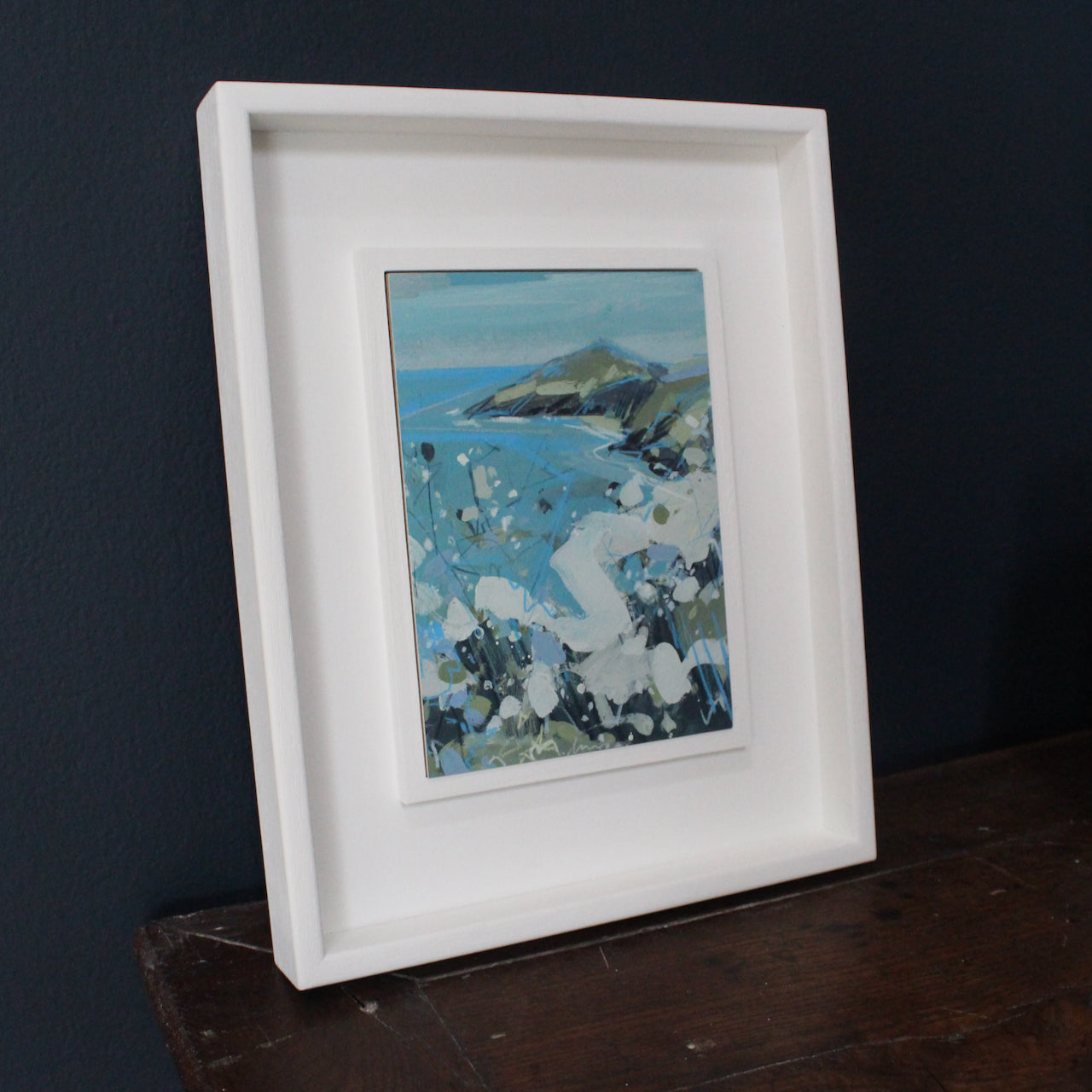 a framed Imogen Bone small painting of Rame Head in Cornwall the sea is turquoise, the peninsula blue and the  hedgerow in the foreground is white and dark green.