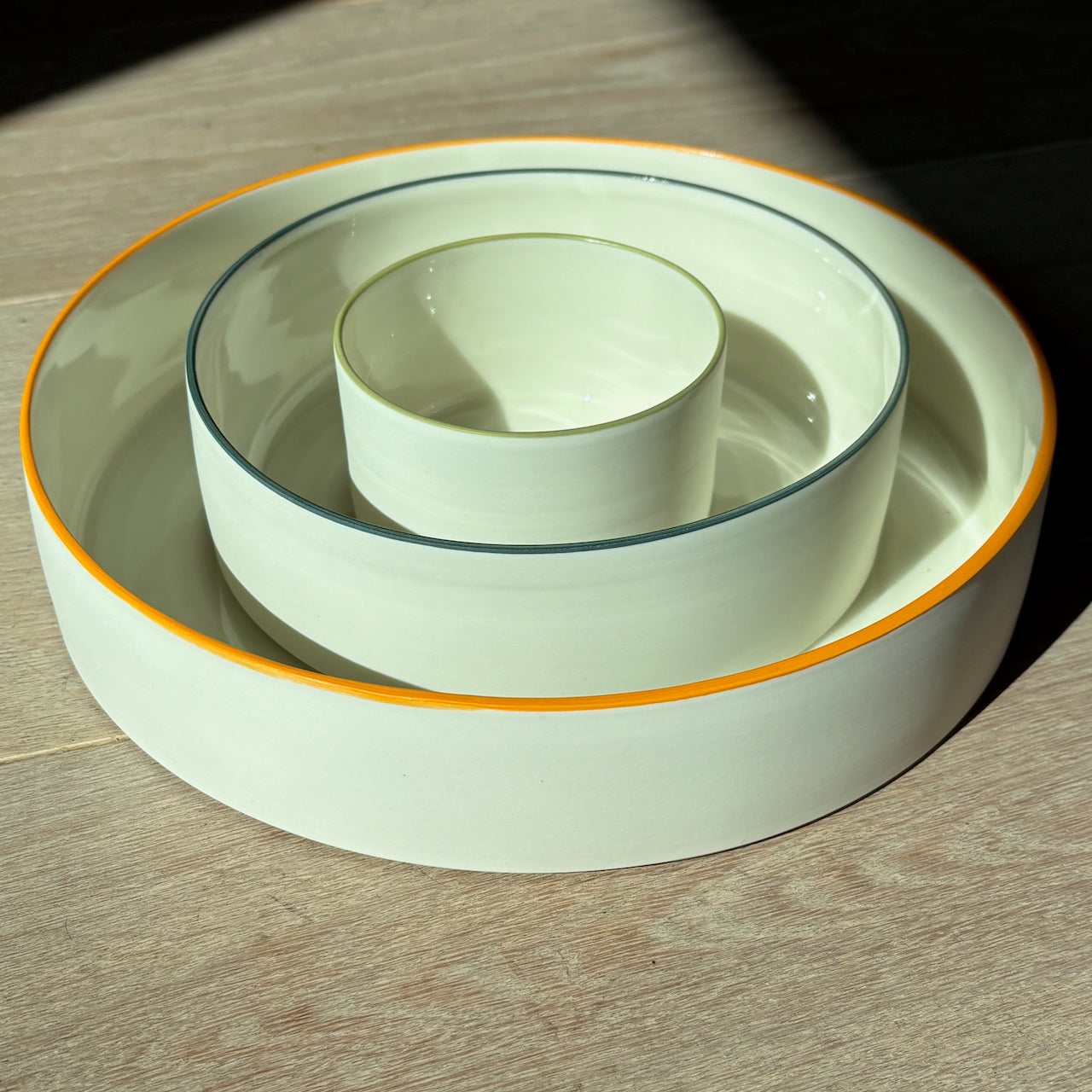 By the Line Pottery, set of three nesting bowls made of porcelain with a coloured rim, the largest has an orange rim, then dark green, then olive