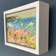 A small oil painting by Jill Hudson of Rame Head in the spring, the headland is green, the sea is bright blue and the foreground shows honeysuckle in pinks and yellows. The painting is framed in a white frame