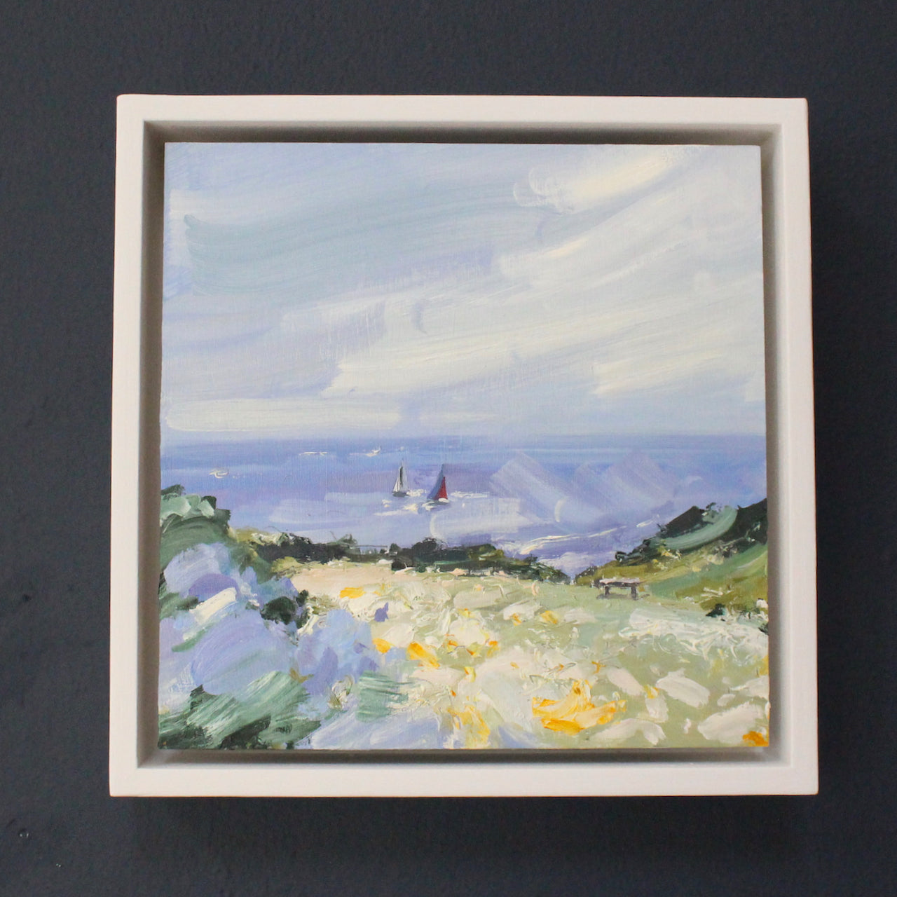 a framed Seascape by artist Jill Hudson of boats sailing in the background and coastal landscape in foreground