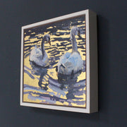 A  framed painting of two swans swimming on gold water by Jill Hudson