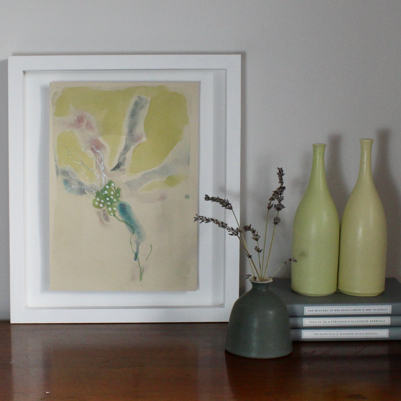 Watercolour of an abstract yellow flower with purple and green details by Tara Leaver  it is in a white frame propped next to two pale yellow ceramic bottles by Lucy Burley
