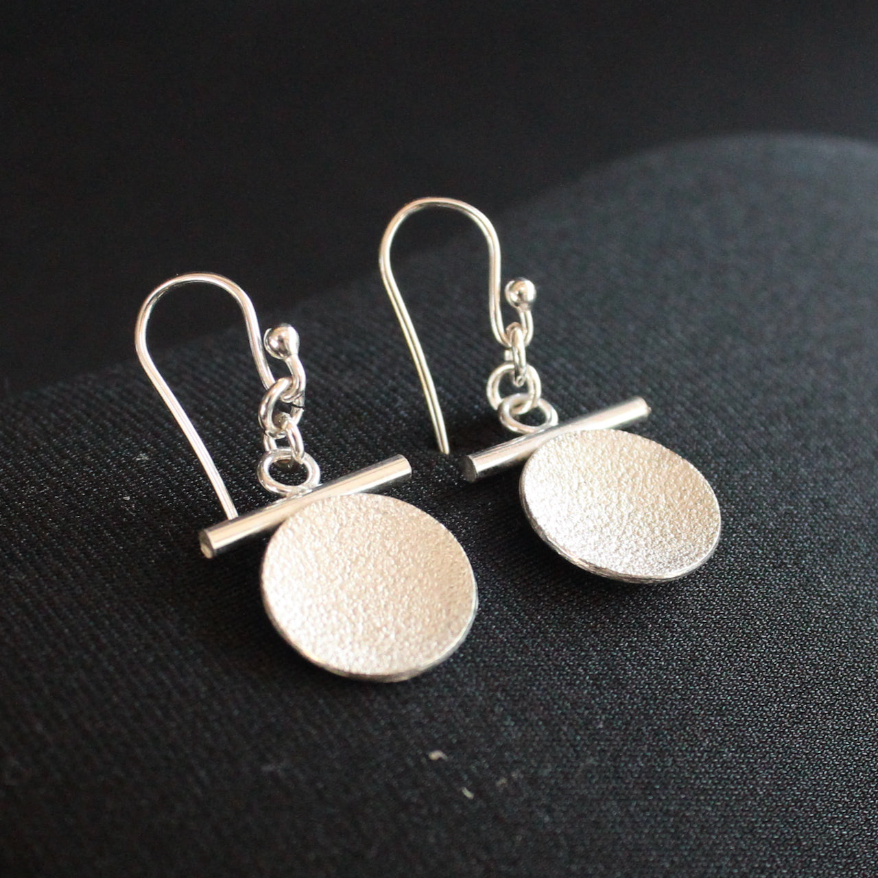 Beverly Bartlett silver drop earrings with a textured silver disc under a silver horizontal bar and silver chain.