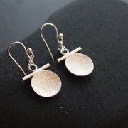 a pair of Beverly Bartlett silver drop earrings with a textured silver disc under a silver horizontal bar and silver chain