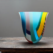 a tall and gently curved multi-coloured glass vase on a wooden table by Ruth Shelley