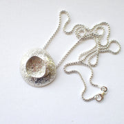 large silver pendant with a delicate circle design on the silver and an indent in the centre it's on a silver chain  and is by jewellery designer Ann Bruford
