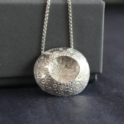 large silver pendant with a delicate circle design on the silver and an indent in the centre it's on a silver chain  and is by jeweller Ann Bruford.