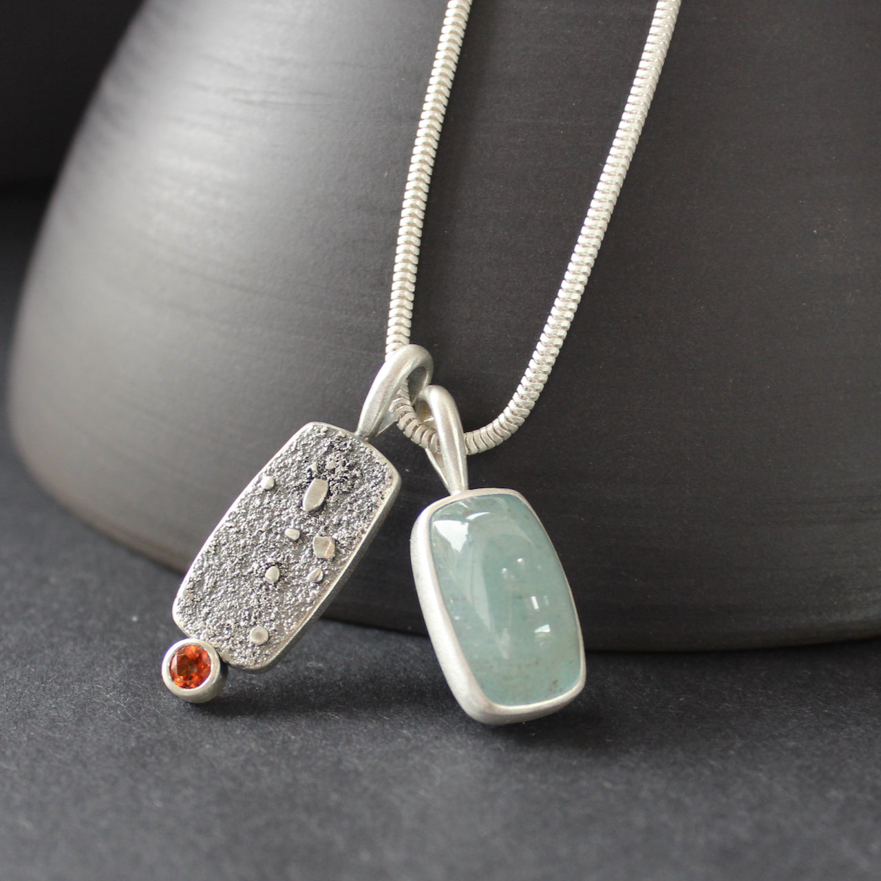 a double silver pendant on a silver chain with one green stone and one small orange topaz it is by  Carin Lindberg.