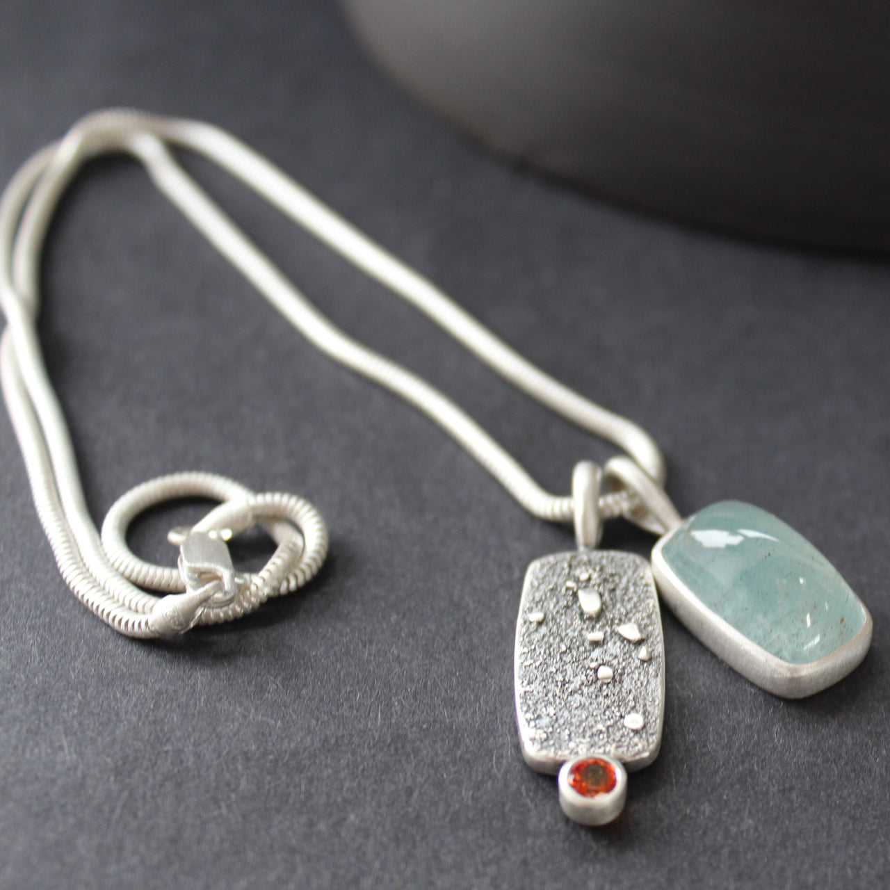 double silver pendant on a silver chain with one green stone and one small orange topaz it is by Cornwall based Swedish jeweller Carin Lindberg