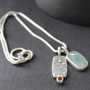 double silver pendant on a silver chain with one green stone and one small orange topaz it is by Cornwall based Swedish jeweller Carin Lindberg