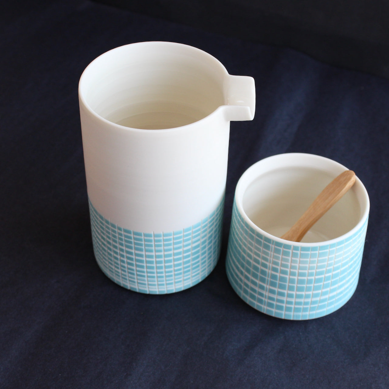 a porcelain pourer and condiment bowl with a wooden spoon by Kathryn Sherriff of By the Line Pottery they are made of porcelain with a blue cross hatch design which covers the whole bowl and the bottom half of the pourer