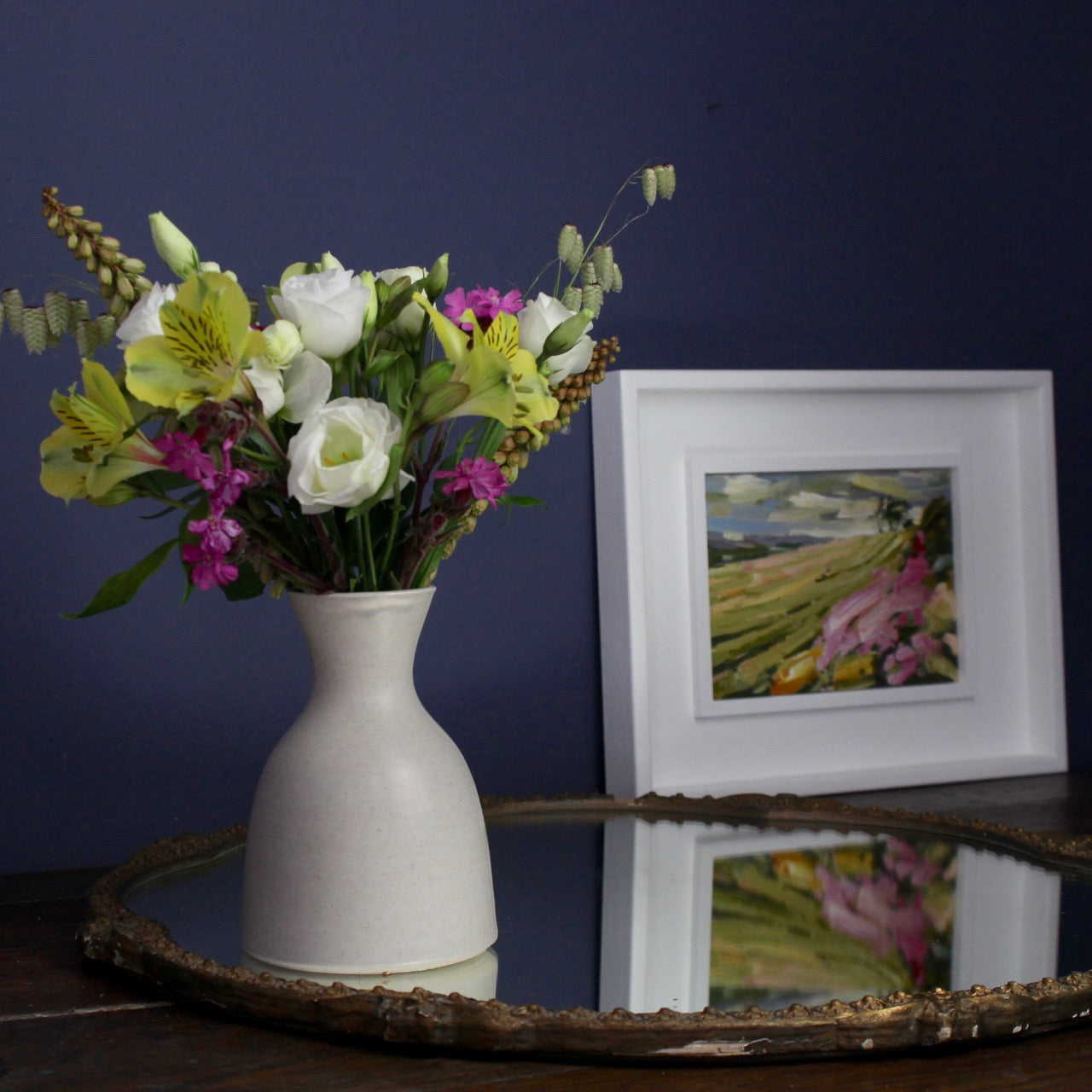 A vase of flowers next to a Jill Hudson framed abstract landscape painting