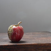 A red ceramic apple with glaze details and a gold stalk by Cornwall ceramicist Remon Jephcott on a wooden table.