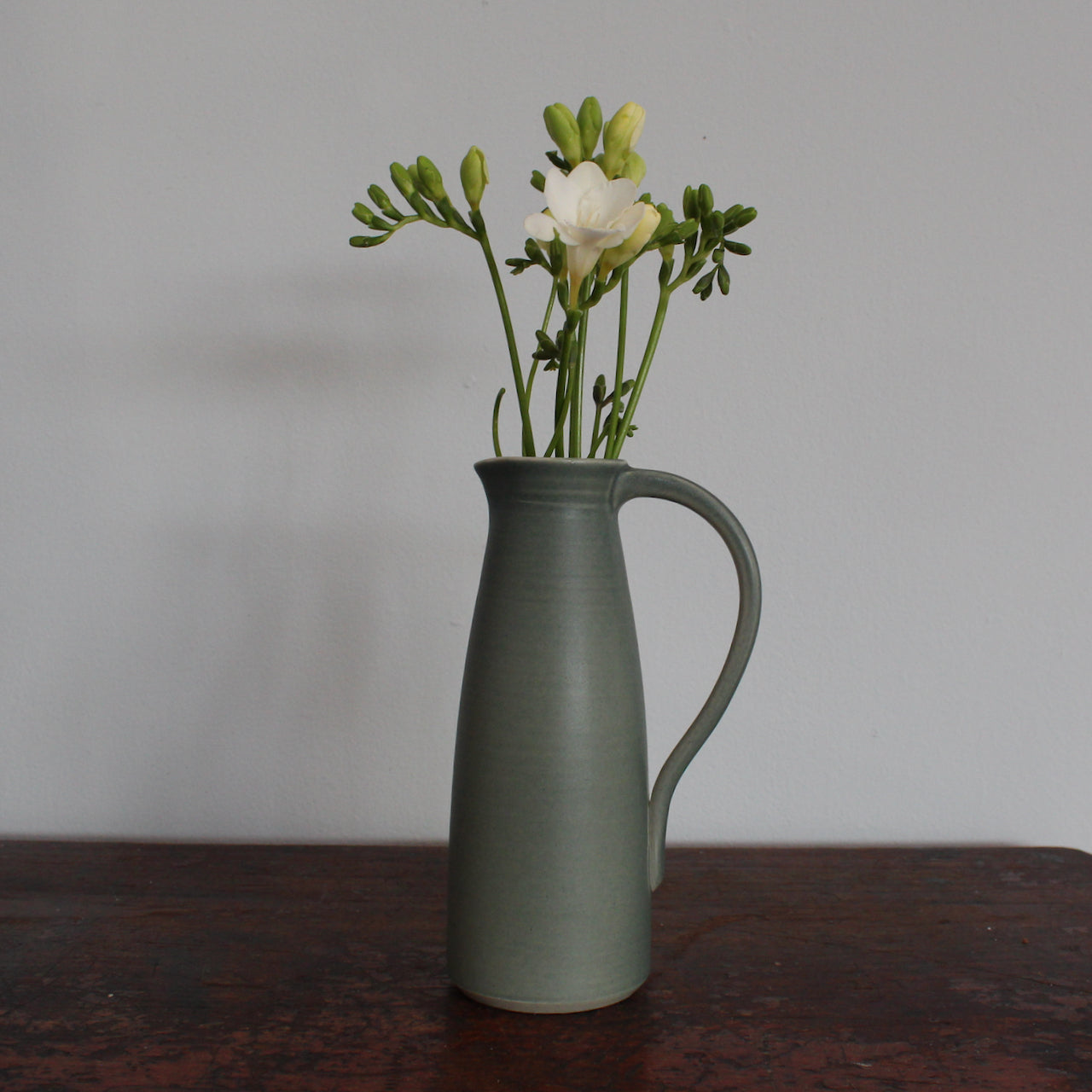 Olive green ceramic jug with handle with flowers in it by Lucy Burley on wooden table at the Byre Gallery