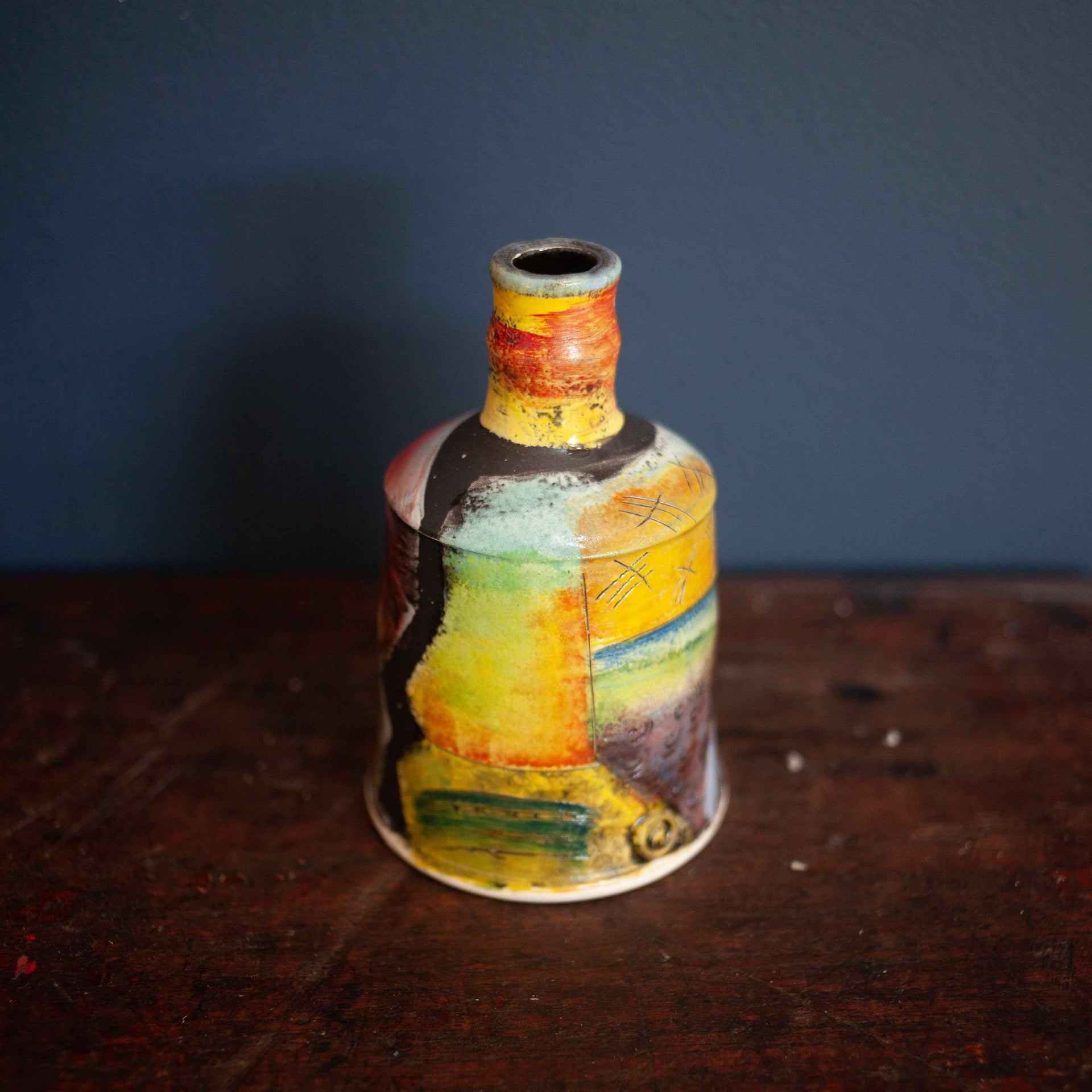 A brightly coloured ceramic bottle by John Pollex on a wooden table with a dark wall