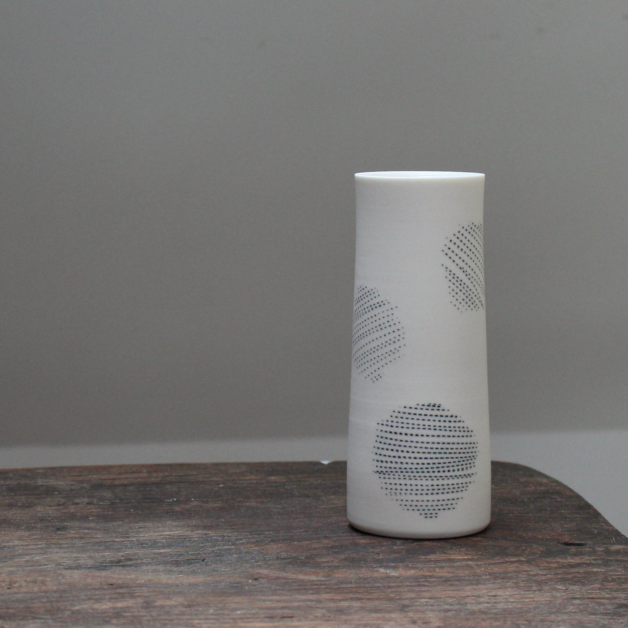 A narrow ceramic white stem vase decorated with abstract circles sitting on a wooden table