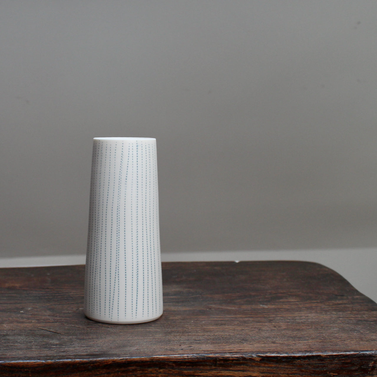 A narrow ceramic white stem vase with vertical blue lines on a wooden table .