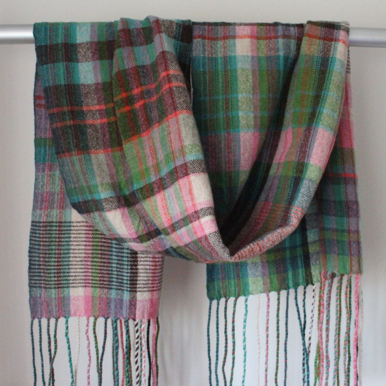 handwoven scarf in shades of pink, green and turquoise hanging over a metal display rail