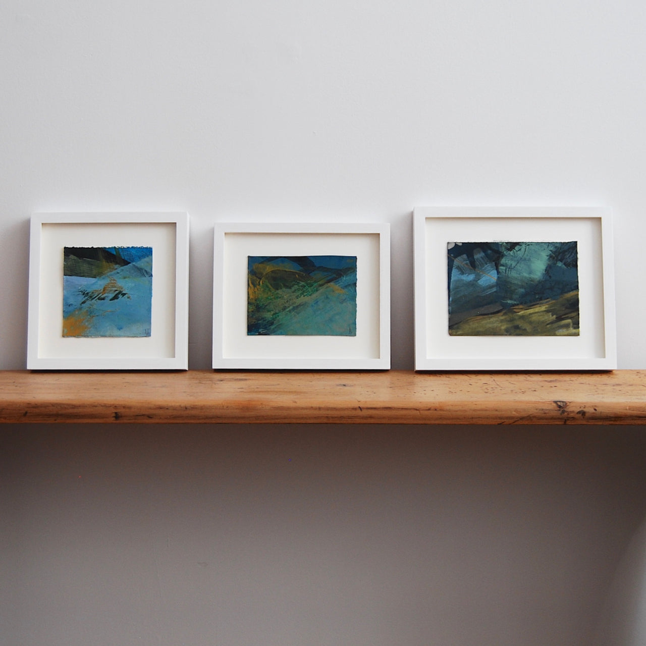 three small abstract landscape paintings by Alice Robinson-Carter in dark blues and yellows they are mounted and framed in white