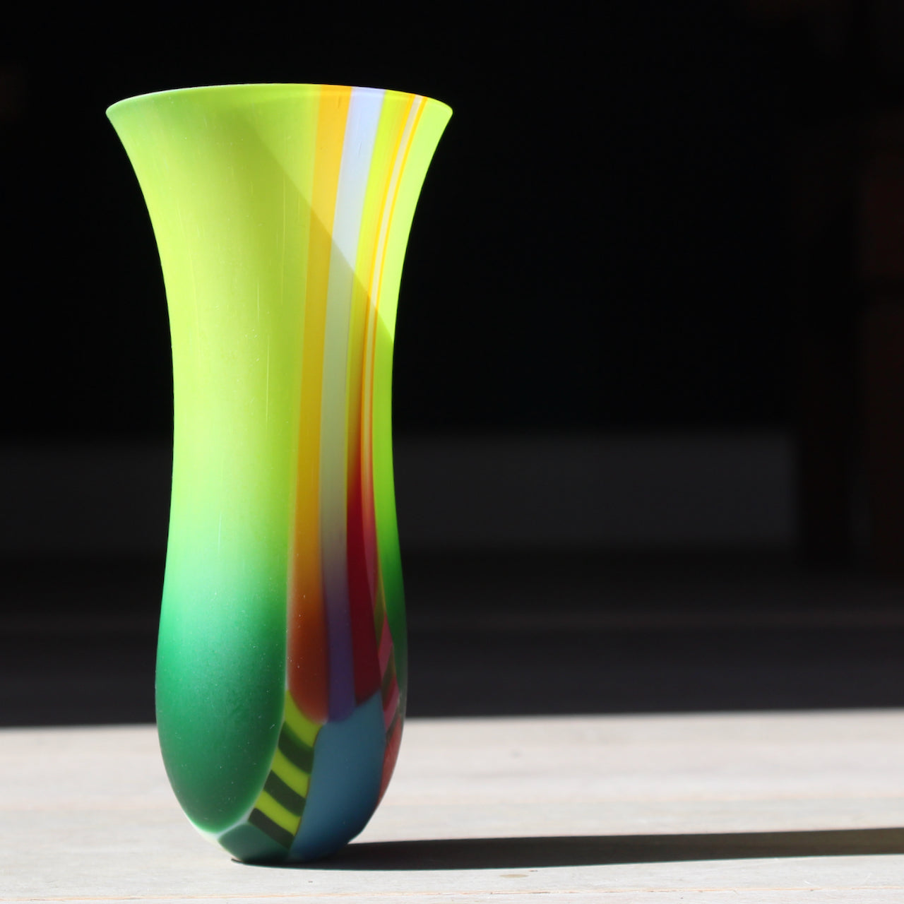 Narrow vibrant coloured glass vase by artist Ruth Shelley in tones of green, yellow, blue and orange.