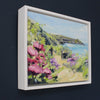a framed Jill Hudson painting of Rame Head in Cornwall with pink flowers in foreground it is on a dark blue wall 