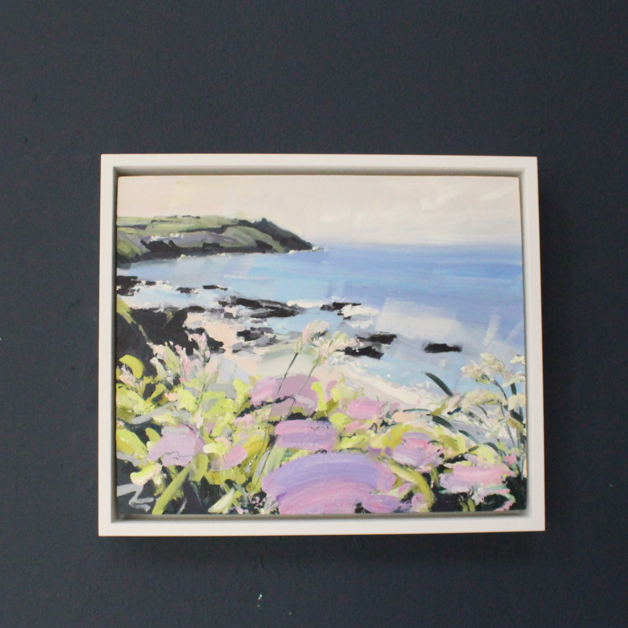a framed Jill Hudson painting of Rame Head in Cornwall with tones of violet, pink and yellows and a blue sea.