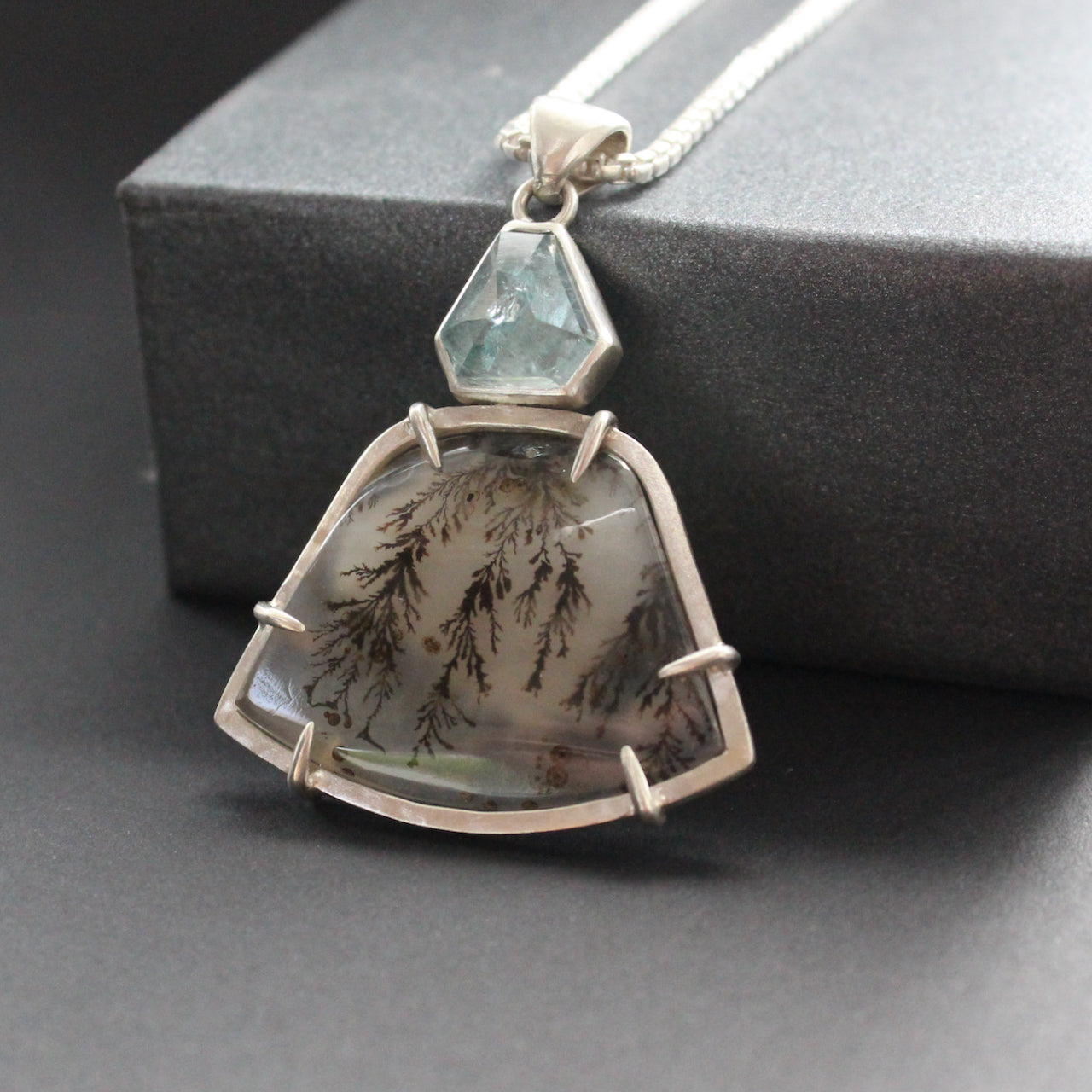 striking pendant by jewellery designer Carin Lindberg featuring small pale blue stone above a grey and detailed agate stone in a silver setting 