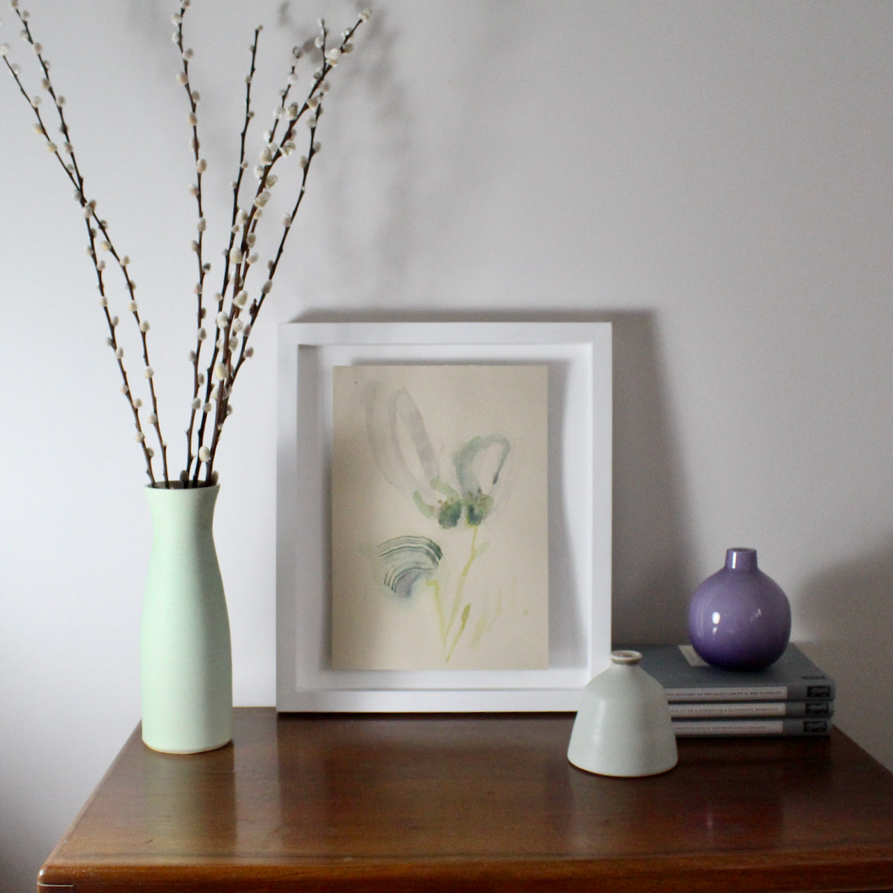 framed watercolour of a flower head and stem by Tara Leaver next to a tall pale green vase with stems in it and a small purple glass pot