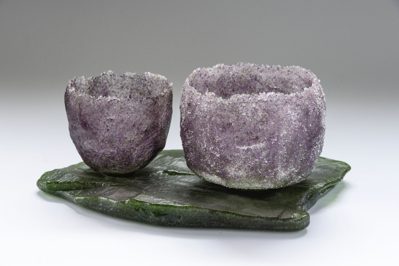 Heavily textured purple glass vessel sitting alongside larger purple vessel on glass slab by artist Caroline Reed