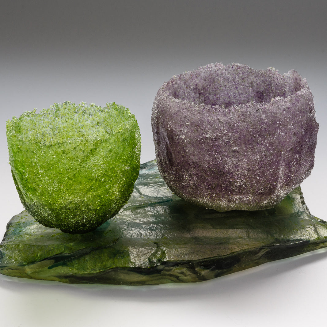 Green heavily textured vessel sitting alongside purple vessel on glass slab by artist Caroline Reed