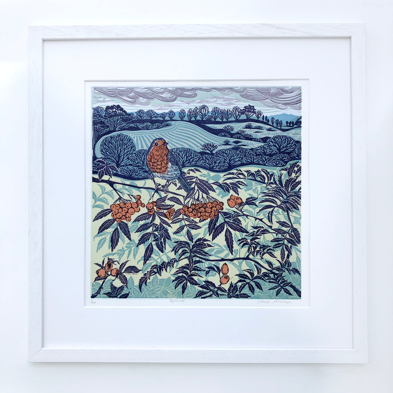 Framed lino print in blue tones with orange of robin sitting on a branch in the foreground and fields and trees in the background by artist Claire Armitage
