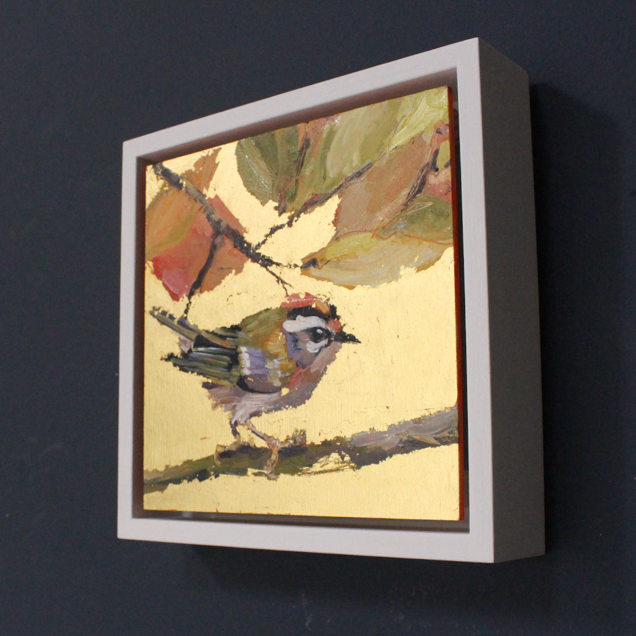 a framed Jill Hudson painting of a firecrest bird on branch in foreground and brown & green leaves.