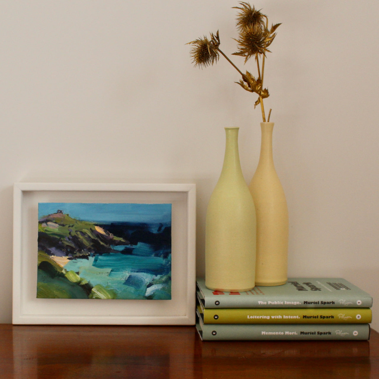 Jill Hudson painting of Rame Head, the headland is in shades of green and dark purple, white hints of golden beach below, the sea is a mix of shades of turquoise underneath a darker sky; the work is framed and propped against a wall next to two pale green  ceramic bottles