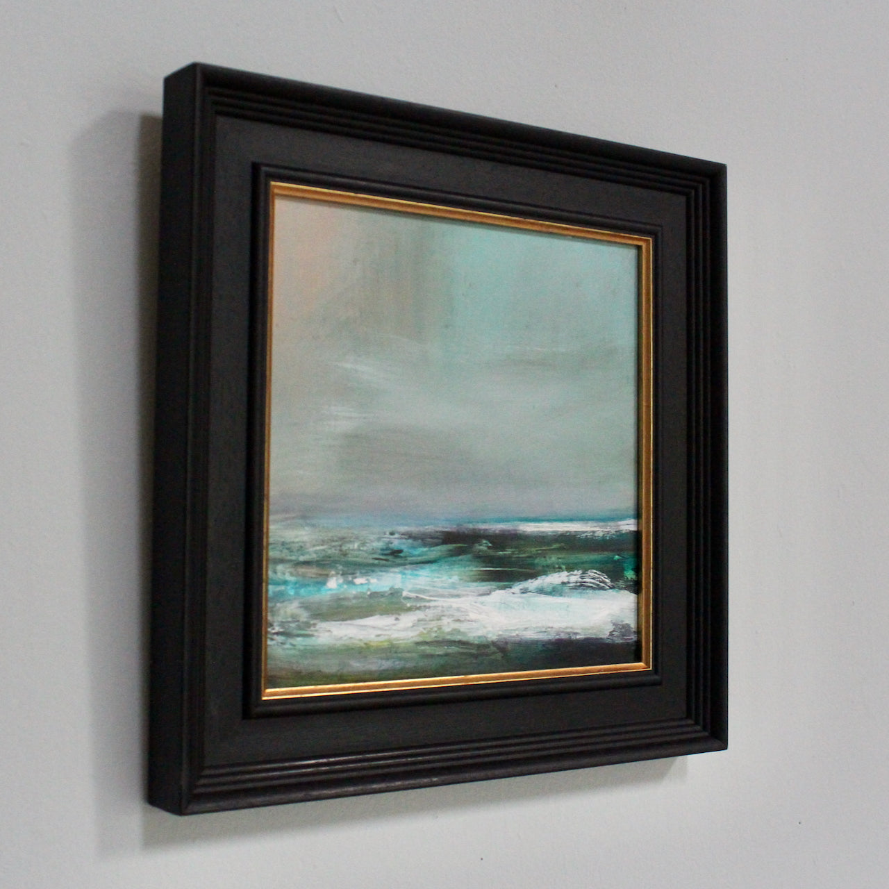 a blue, green and white sea under a grey sky in a painting by UK artist Claire Howlett framed in a black frame.