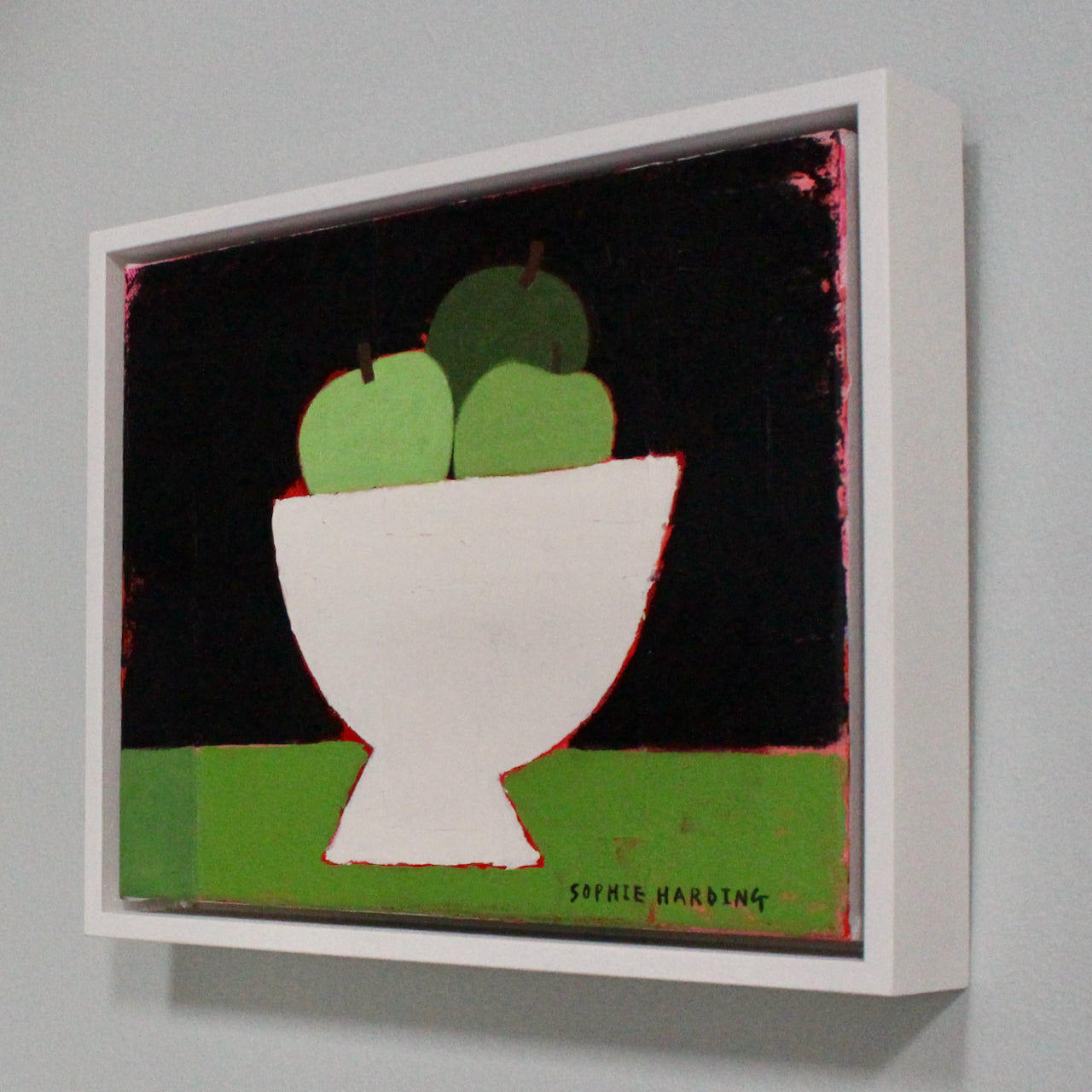 a framed Sophie Harding painting of three green apples in a white bowl on a green coloured table.