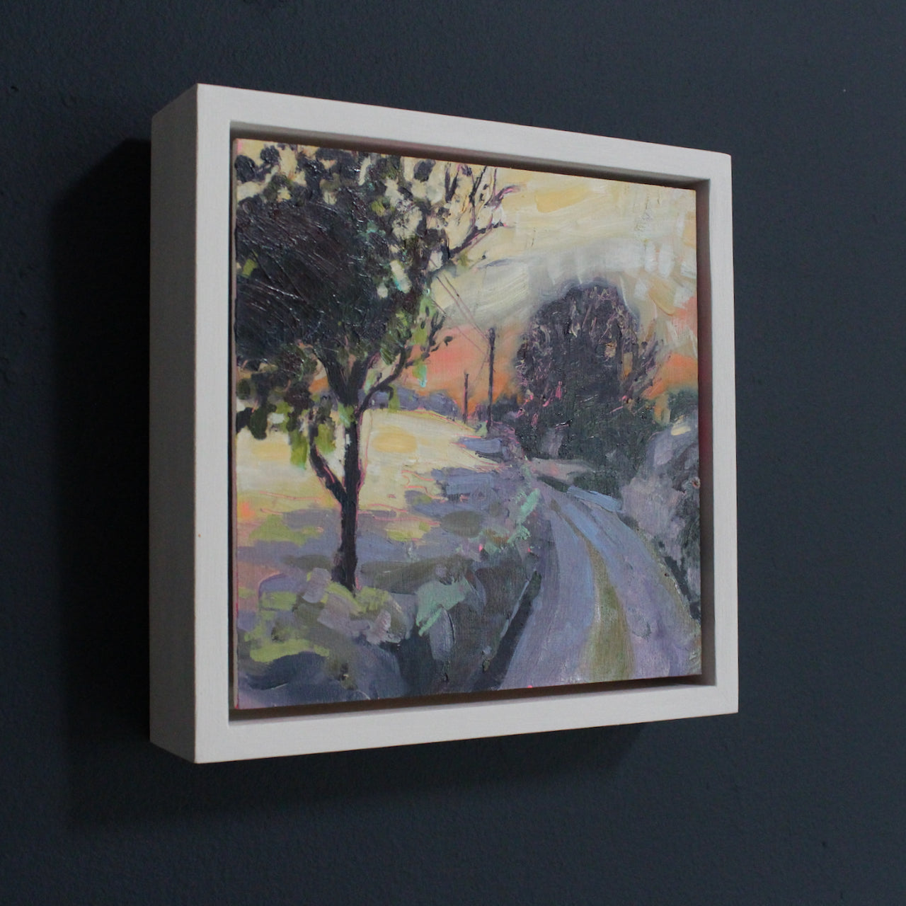 a framed landscape painting by Jill Hudson of a country lane at sunset, an orange sky, dark trees and hints of purple on the road and hedgerows.