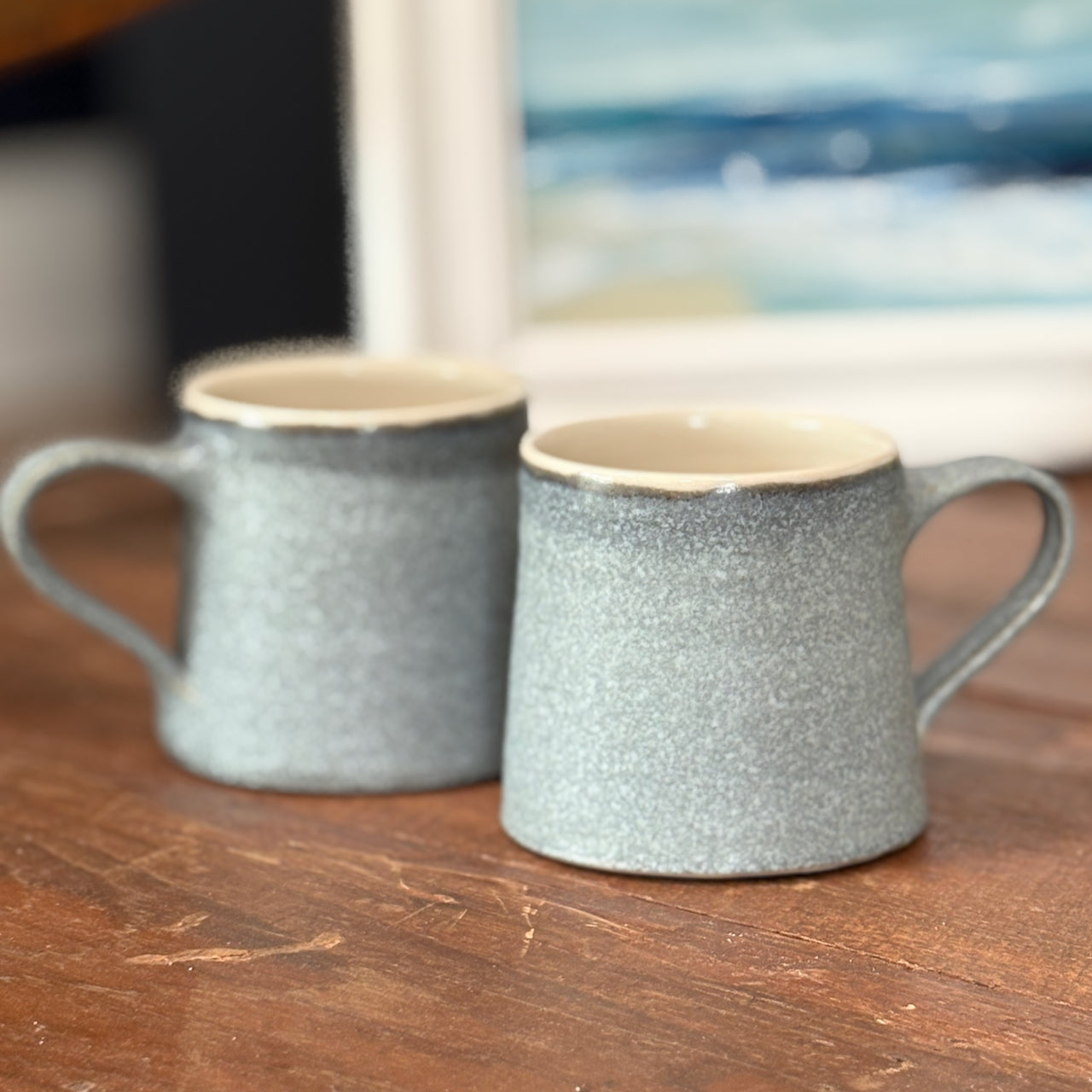EOT ceramics pair of large blue grey mugs on a wooden table with a painting of seascape just visible in the background 