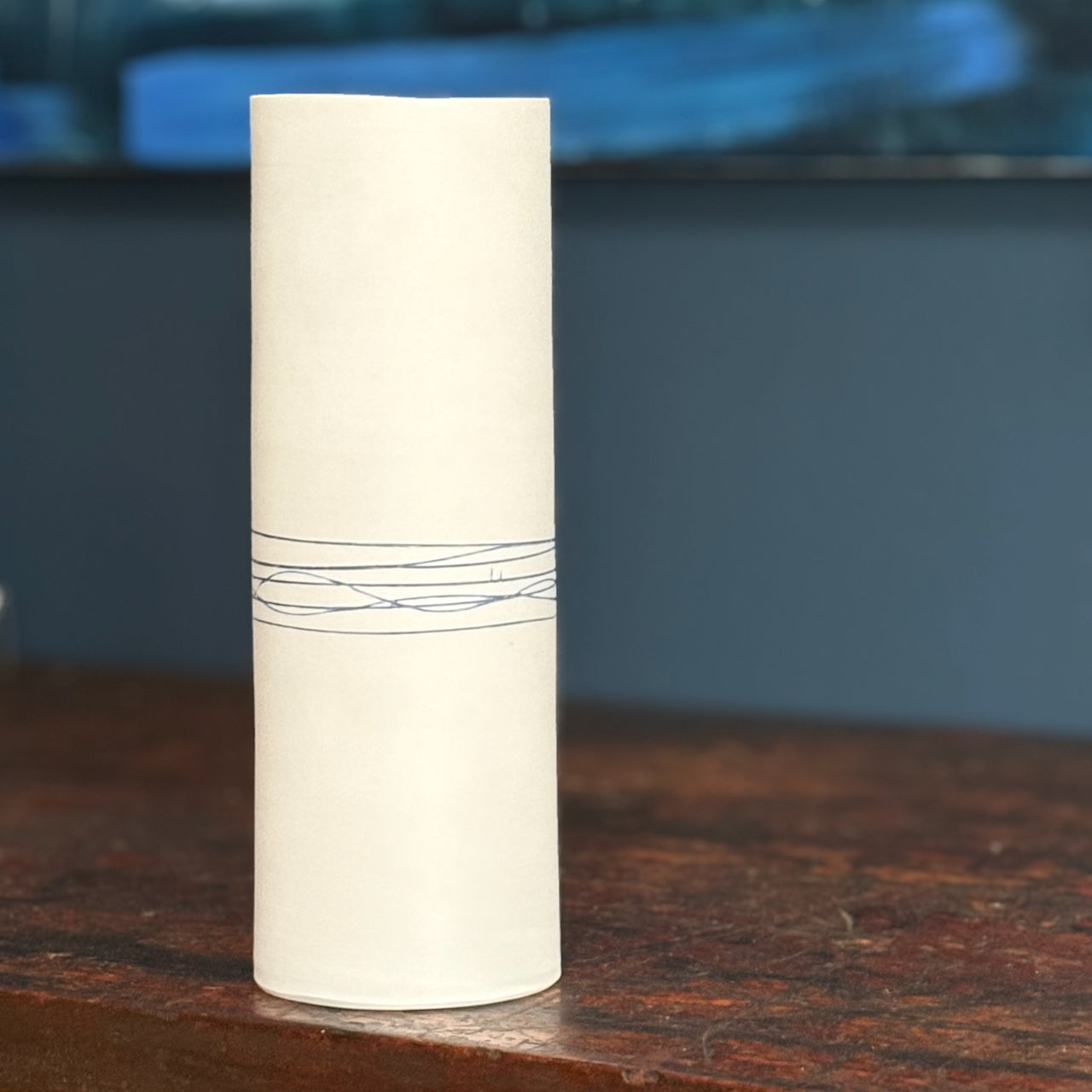 Cylindrical white vase with blue lines on a wooden surface with a blurred blue background by ceramic artist Liz O'Dwyer
