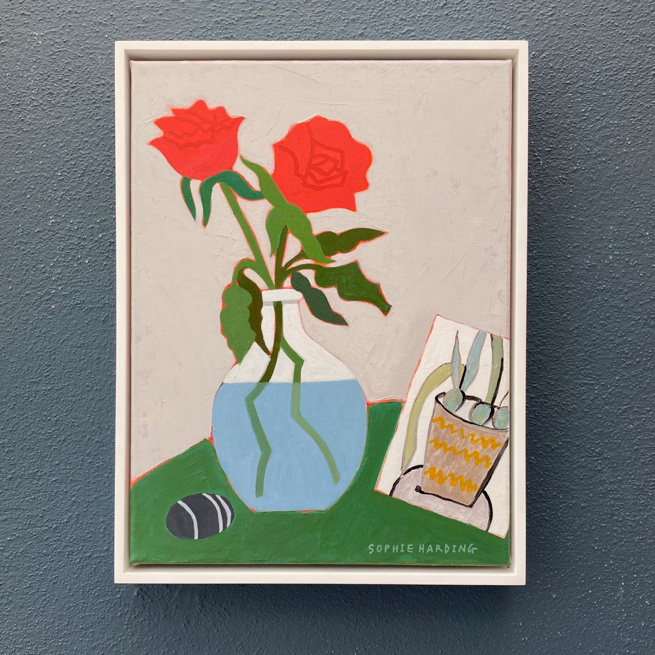 Framed artwork of two red roses in a blue vase on a green surface with a small bag, signed 'Sophie Harding'.