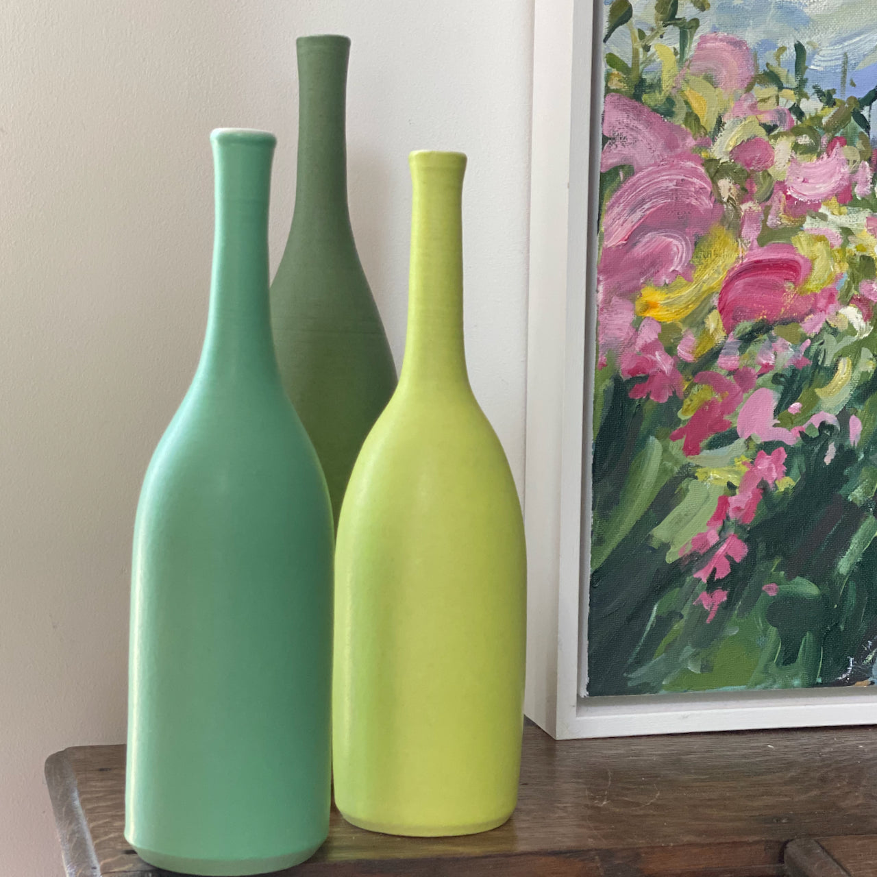 three ceramic bottles in shades of green by Lucy Burley standing next to corner of oil painting showing a pink and yellow hedgerow 