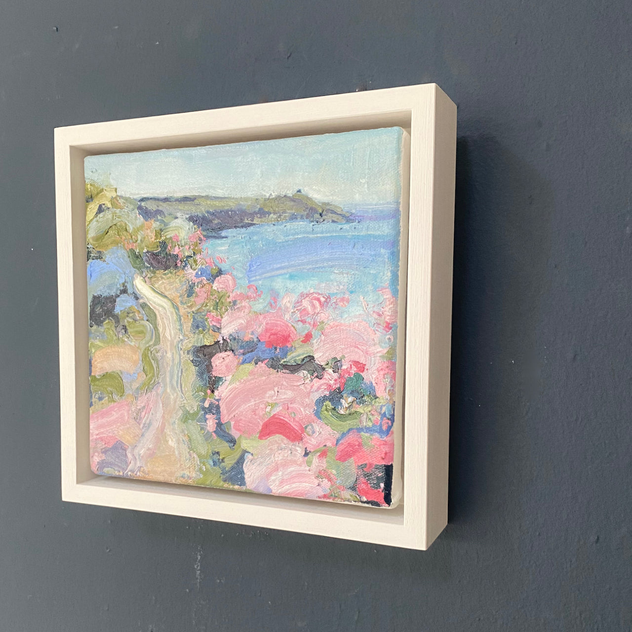 a framed Jill Hudson oli painting of Rame Head in south east cornwall, the headland is dark green with a pale blue and purple sea and the coast path  features pink, white and blue wildflowers.
