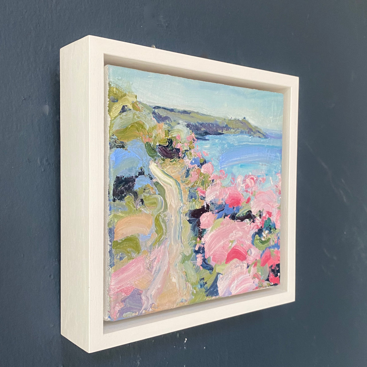 In a deep white frame this is a Jill Hudson oli painting of Rame Head in south east cornwall, the headland is dark green with a pale blue and purple sea and the coast path  features pink, white and blue flowers