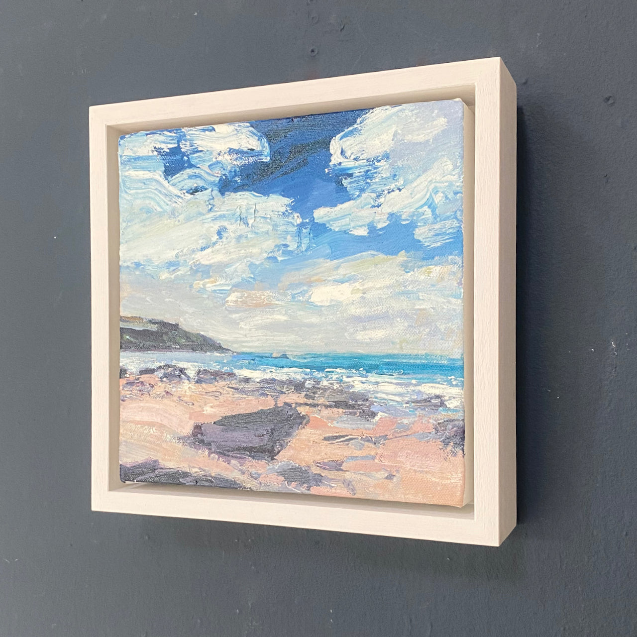 a framed Jill Hudson oil painting of a beach on the Rame peninsula in south east Cornwall, a blue sky with thick white clouds is over a turquoise sea and pink toned sand with grey and purple rocks