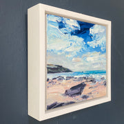 Framed in a deep white wooden frame is a Jill Hudson oil painting of a beach on the Rame peninsula in south east Cornwall, a blue sky with thick white clouds is over a green sea and pink toned sand and black rocks