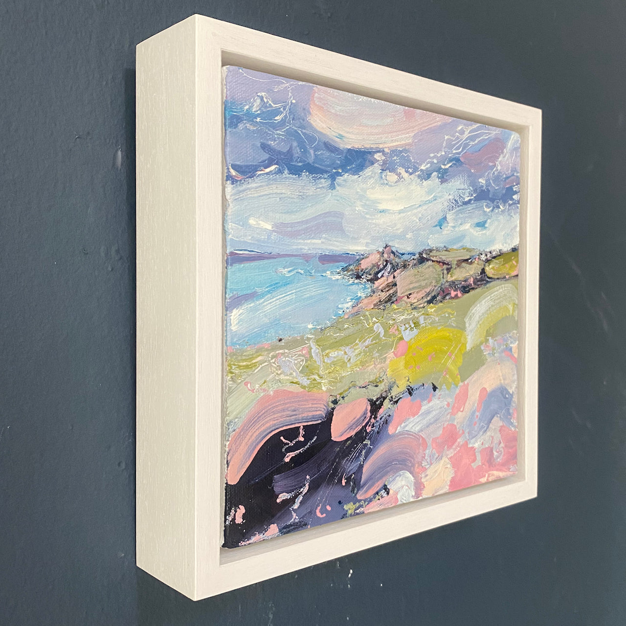 a framed colourful Jill Hudson oil painting of Rame Head in south east Cornwall: the headland is green and pink under a blue and white sky and the foreground shows yellow and pink wildflowers.