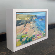 a small Jill Hudson painting of Rame Head in Cornwall, the headland is green with a golden beach framed by summer wildflowers; the painting is framed.