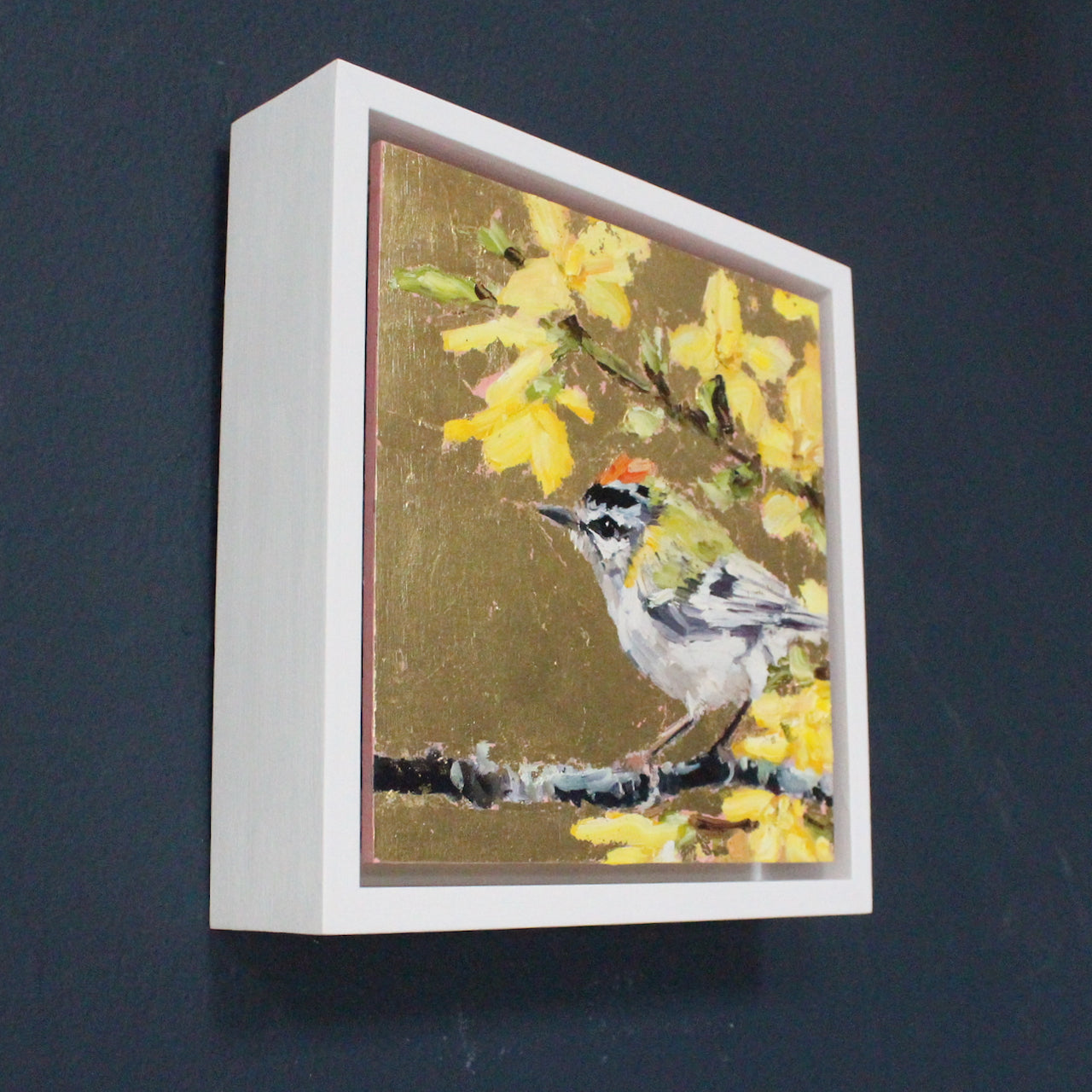 Framed square painting with gold lustre background, firecrest sitting on branch in foregound and yellow/cream blossom by artist Jill Hudson