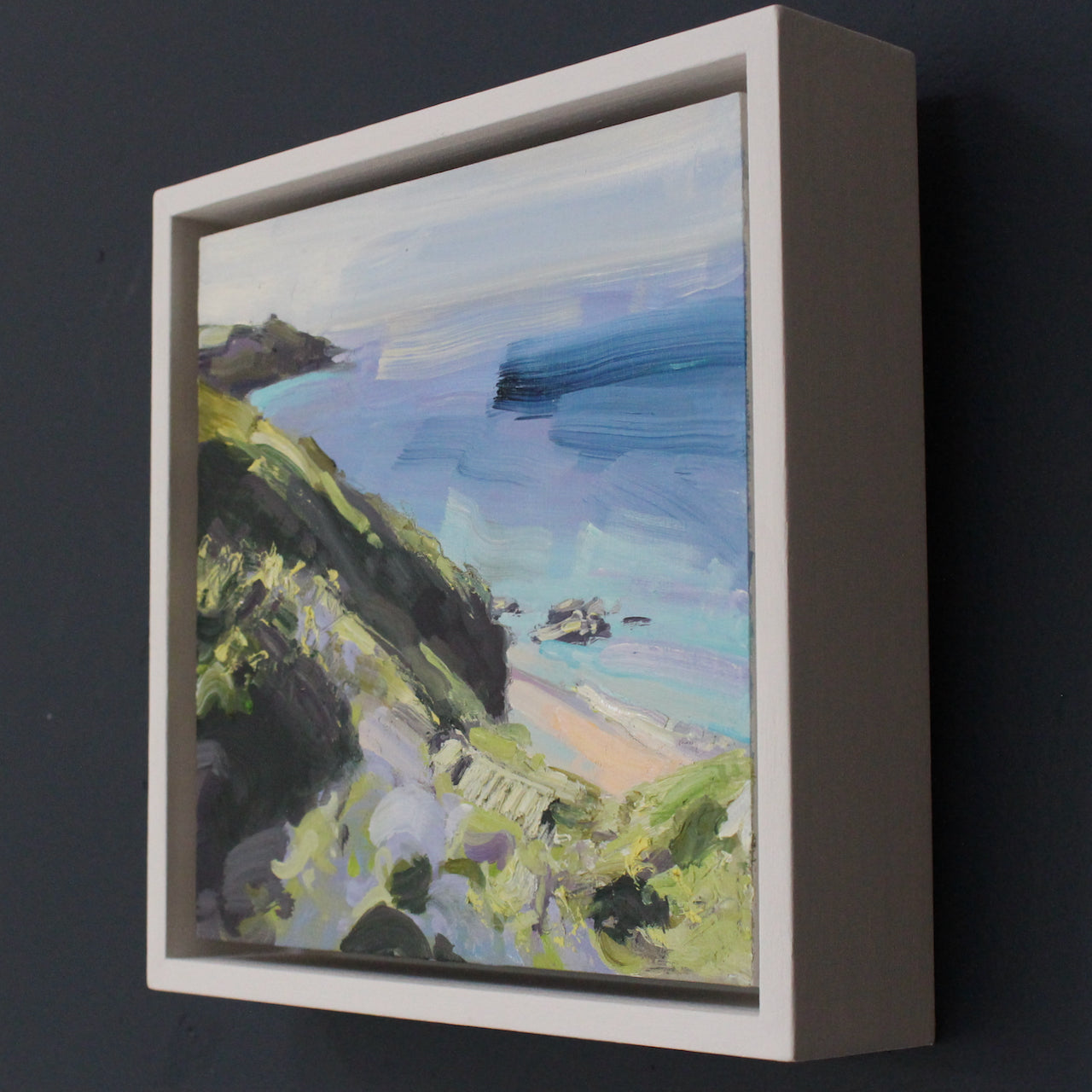 a framed Jill Hudson painting of Rame Head in Cornwall in pastel tones with dark green cliffs 